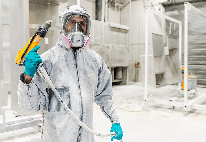 Crestline worker wearing full protective gear and respirator holds a spray gun inside the paint booth, preparing to apply a powder coat finish to an ambulance module