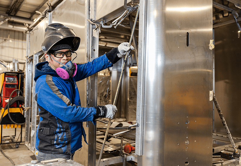 Worker wearing protective gear and a respirator works on the aluminum frame of an ambulance module inside the Crestline manufacturing facility