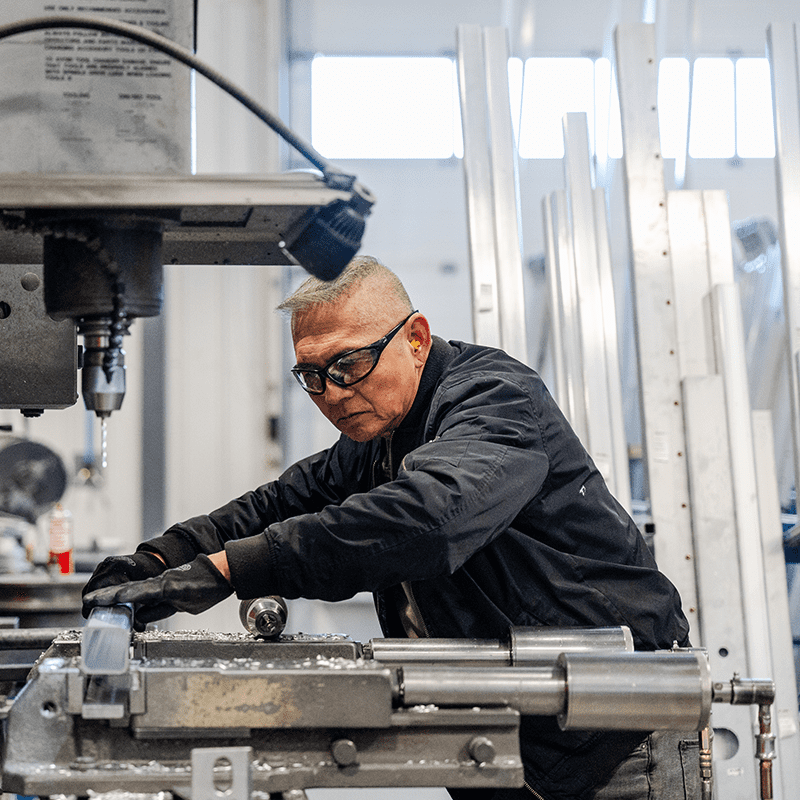 A Crestline factory worker wearing safety glasses, ear protection, and gloves operates a milling machine to shape a metal part for the ambulance module.