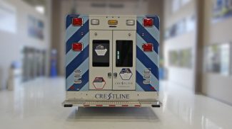 Rear view of an ambulance with blue diagonal stripe graphics, dual rear access doors, red and amber emergency lights, and Crestline branding