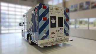 Rear side view of an ambulance inside a showroom, featuring bold blue graphics, rear double doors with safety decals, red and amber emergency lights, and Crestline branding