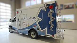 Side angle of an ambulance, featuring a large blue Star of Life graphic with caduceus symbol, side access doors, emergency lighting, and Crestline branding, displayed inside a showroom