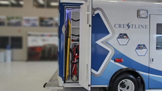Exterior side compartment of a Crestline ambulance open, showing organized storage with a yellow backboard and red stretcher securely mounted inside