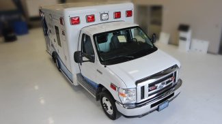 Overhead view of a white Crestline Ford E-350 ambulance with red emergency lights, parked indoors on a clean white floor