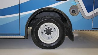 Close-up of a Crestline ambulance rear wheel with white rim, black tire, and visible fuel cap labeled 'Gasoline Only' against the vehicle’s blue and white exterior design