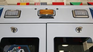 Close-up of the rear upper section of a Crestline ambulance showing two rear door windows, chrome latches, LED scene lights, and an amber warning light with an integrated rearview camera