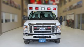 Front view of a white Crestline ambulance with red and white emergency lights, displayed indoors on a polished white floor