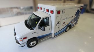 Overhead angled view of a white Crestline Ford ambulance with red emergency lights and blue side graphics, parked indoors on a polished white floor