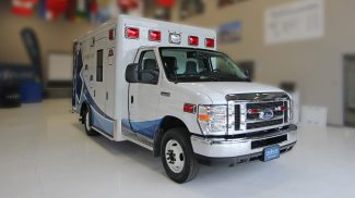 Side angle view of a white Crestline Ford ambulance with red emergency lights, chrome grille, and blue wave graphics, displayed indoors on a polished floor
