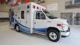 Side view of a white Crestline Ford E-350 ambulance with blue and red graphics, chrome grille, and red emergency lights, parked indoors on a polished floor