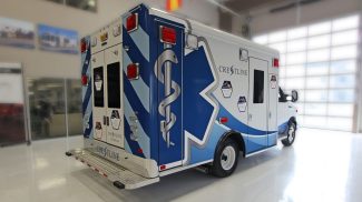 Rear side view of a Crestline ambulance parked indoors, featuring blue and white graphics with the Star of Life emblem, red emergency lights, and rear access doors
