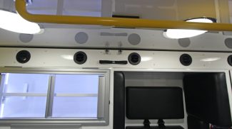 Interior of a Crestline ambulance showing overhead yellow safety rail, bright LED dome lights, circular air vents, and black padded seating beside storage cabinets with sliding glass doors
