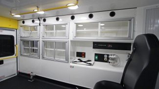 Interior of a Crestline ambulance featuring medical cabinets with sliding glass doors, overhead lighting, wall-mounted controls, and a black attendant seat