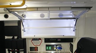 Crestline ambulance interior cabinet with upward-opening glass door, illuminated storage space, and nearby control panel with medical equipment and outlets