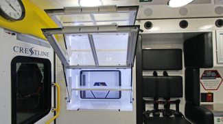 Interior of a Crestline ambulance showing illuminated overhead aluminum cabinet with upward-opening glass doors open and seating area with safety harnesses