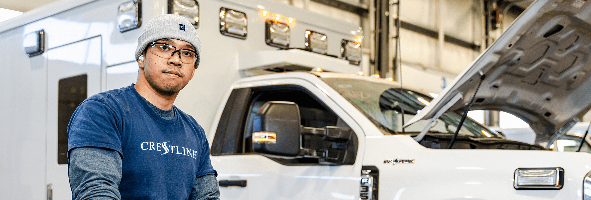 Crestline team member wearing safety glasses and a beanie works in the production facility beside a white ambulance with its hood open