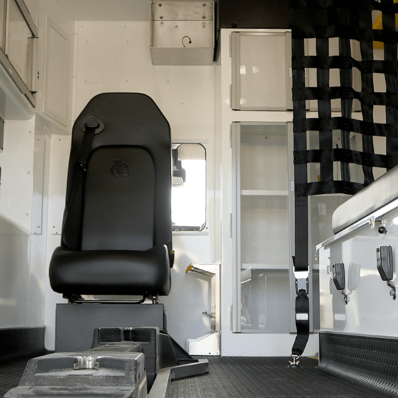 Interior of a Crestline ambulance showing a black attendant seat with seatbelt, storage cabinets, equipment mounts, and safety net separating compartments.