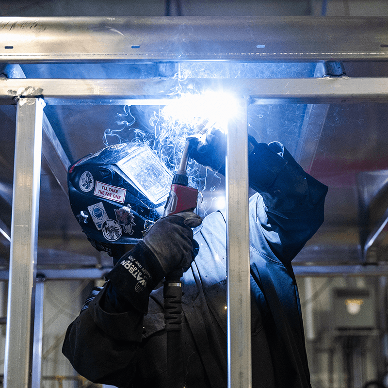 Crestline worker wearing protective gear and helmet works on an aluminum ambulance frame, with bright sparks and smoke visible from the welding torch