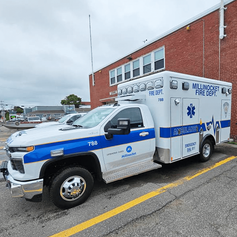 Blue and white Millinocket Fire Department Crestline ambulance parked outside a brick fire station building on a cloudy day