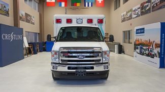Front view of a Crestline CCL 166 ambulance built on a Ford chassis, displayed inside the Crestline manufacturing facility with branding and international flags in the background