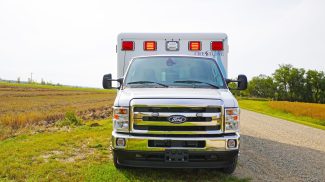 Front view of a Crestline ambulance with red emergency lights on, parked on a gravel road beside an open rural field