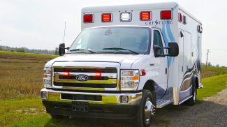 Front view of a Crestline ambulance with emergency lights illuminated, parked on a rural roadside surrounded by open fields