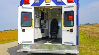 Rear view of a Crestline ambulance with its back doors open, showcasing the clean and organized patient care interior against a rural field backdrop