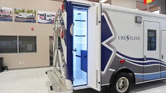 Rear-side view of a Crestline ambulance with an open exterior compartment illuminated in blue light, showing interior shelving and storage space