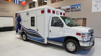 Crestline CCL 166 ambulance shown in a showroom, featuring blue and gray wave graphics along the sides and a large Star of Life design on the rear module