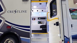 Open side door of a Crestline ambulance revealing the illuminated interior with black seating, control panels, and diamond plate entry steps