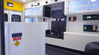 Interior of a Crestline ambulance featuring black seating with seatbelts, upper and lower storage cabinets, oxygen access panel, and control switches