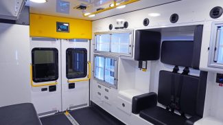 Crestline interior view of an ambulance showing rear entry doors with yellow grab handles, black seating with seatbelts, overhead lighting, and illuminated storage cabinets