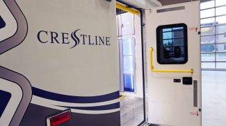 Side view of a Crestline ambulance with the entry door open, revealing bright yellow grab handles, diamond plate steps, and illuminated interior cabinets