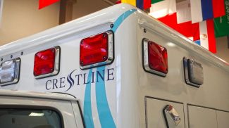 Close-up of the upper rear corner of a Crestline ambulance featuring red and clear warning lights, smooth body lines with blue accent graphics, and the Crestline logo, captured indoors beneath a display of international flags