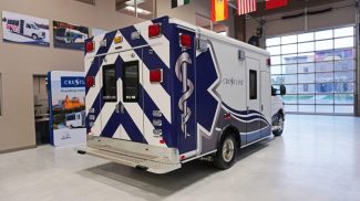 Rear and side view of a Crestline CCL 166 ambulance featuring a blue and white chevron design with the Star of Life graphic, photographed inside the Crestline manufacturing facility showroom