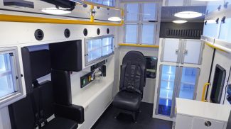 Wide interior view of a Crestline ambulance featuring black seating with seatbelts, upper and lower storage cabinets, medical equipment controls, and bright yellow grab handles