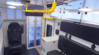Wide view of a Crestline ambulance interior featuring black seating with safety harnesses, illuminated storage cabinets, yellow grab handles, and overhead lighting