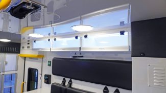 Overhead view inside a Crestline ambulance showing upper storage cabinets with clear doors, ceiling lighting, yellow grab handles, and a black bench seat below