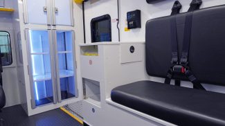 Crestline ambulance interior featuring a black bench seat with seatbelts, illuminated storage cabinets, a biohazard waste disposal unit, and a digital control panel