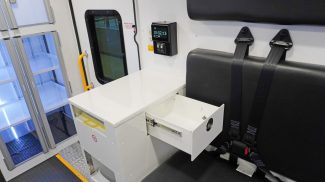 Crestline ambulance interior featuring a black bench seat with seatbelts, an open storage drawer above a biohazard waste unit, and illuminated supply cabinets near the side door