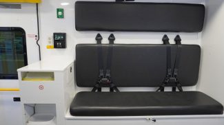 Crestline ambulance interior featuring a black bench seat with integrated seatbelts, wall-mounted backrest cushions, a biohazard waste disposal unit, and control panel with power outlets