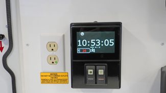 Close-up view of a Crestline ambulance interior wall panel featuring a digital control screen displaying the time and battery status beside a dual 120V power outlet with a caution label