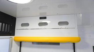 Closed overhead storage compartment inside a Crestline ambulance with oval cutouts, a bright yellow grab handle, and a ceiling light above
