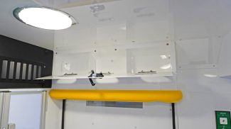 Overhead storage compartment inside a Crestline ambulance with an open latch door, bright ceiling light, yellow grab handle, and a nearby oxygen outlet