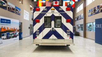 Rear exterior view of a Crestline CCL 166 ambulance featuring a blue and white chevron design, rear entry doors with Crestline branding, and a diamond plate step bumper inside the Crestline manufacturing facility