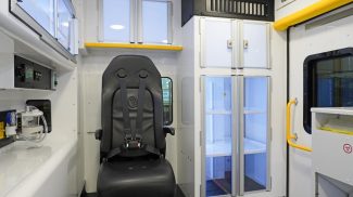 Interior view of a Crestline ambulance module featuring a black attendant seat with a four-point harness, illuminated storage cabinets with clear doors, and bright yellow grab handles near the side entry door