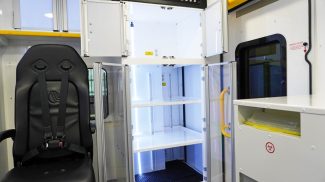 Interior view of a Crestline ambulance module featuring a black crew seat with a four-point harness beside a brightly lit medical supply cabinet with clear doors and adjustable shelves