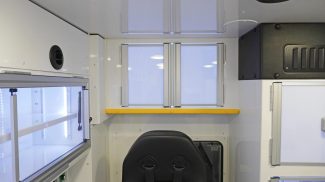 Interior view of a Crestline ambulance showing closed upper storage cabinet doors above the attendant seat, with a yellow safety rail and adjacent LED-lit aluminum cabinets along the side wall