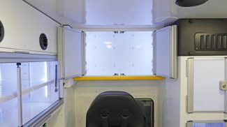 Interior view of a Crestline ambulance showing an open upper storage cabinet with dual doors above the attendant seat, featuring bright interior lighting, a yellow safety rail, and adjacent aluminum-framed cabinets