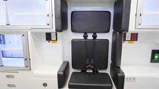 Rear-facing attendant seat inside a Crestline ambulance, featuring integrated black padding, a 4-point seatbelt, and surrounding aluminum storage cabinets with electrical outlets and medical control panels nearby